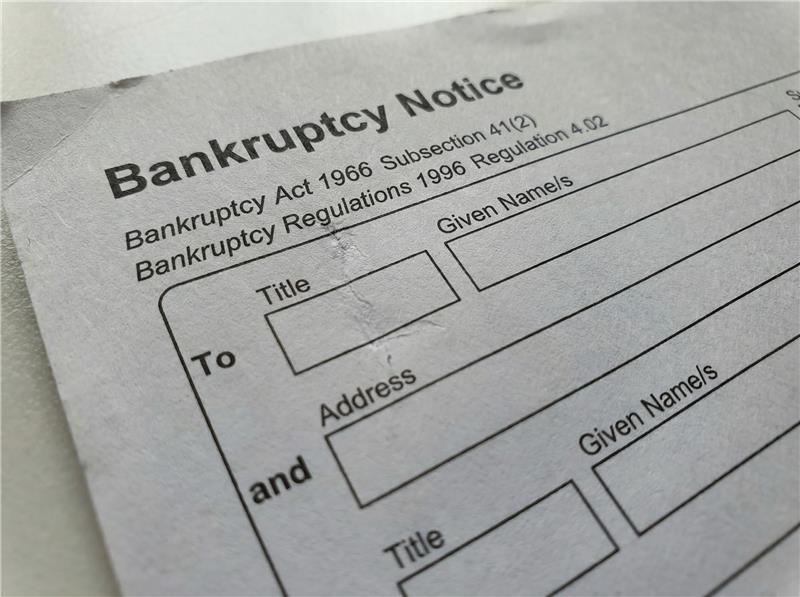 A person standing indoors looks out through large glass windows toward a parked hatchback car with a colorful geometric wrap. The car is in an outdoor lot surrounded by trees and a modern two‑story office building.